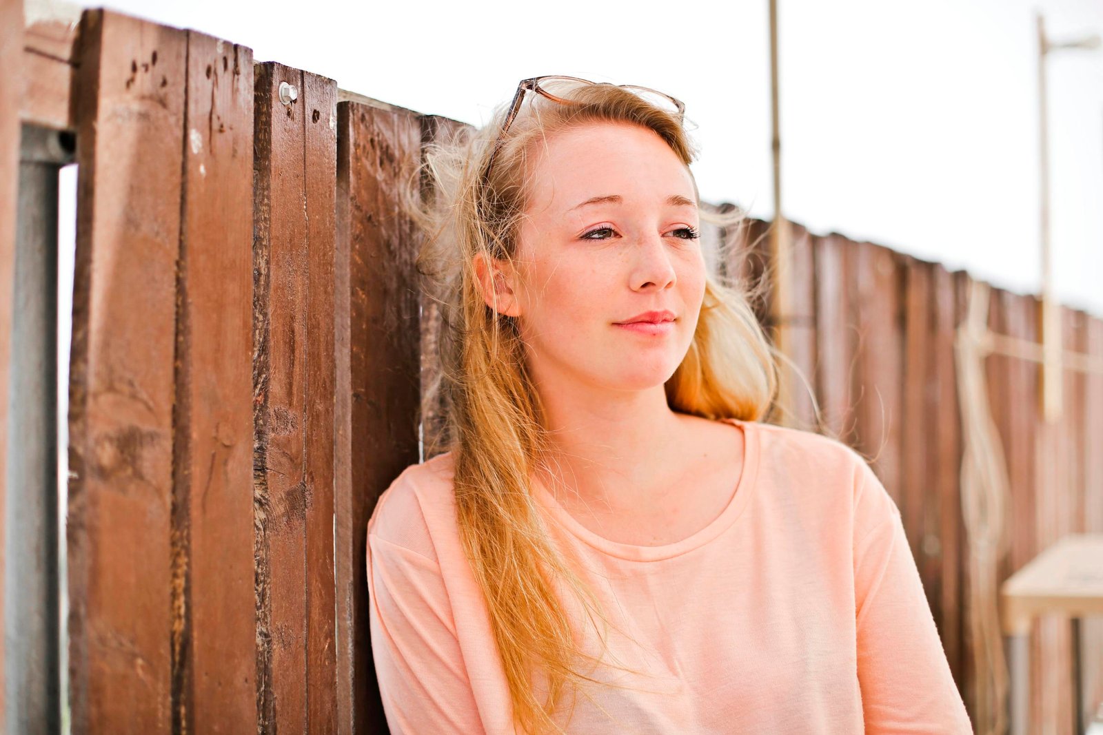 Portrait of a young woman outdoors leaning against a wooden fence, looking thoughtful and relaxed.